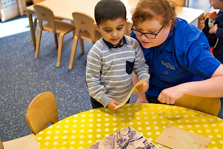 An early years practitioner is looking at a table with a young boy in a classroom. The table is covered with sand and pictures of dinosaur bones and fossils.