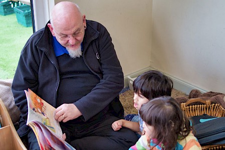 An early years practitioner and two young girls. He is reading them a story in a cosy corner of the nursery.