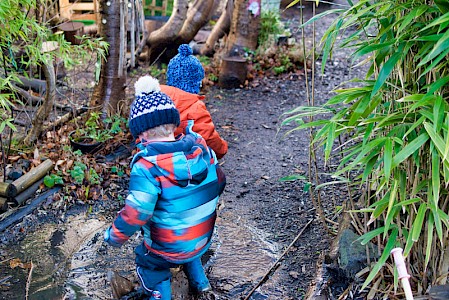 Two young boys in wellies and winter clothing jumping in a puddle in the nursery’s garden.