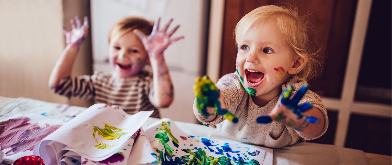 Two smiling young children are finger painting. They have their paint covered hands showing to the camera.